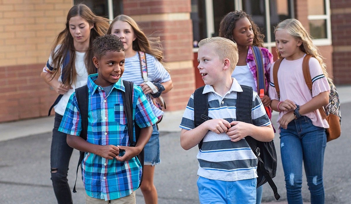 Children wearing backpacks walk and talk in front of a school building